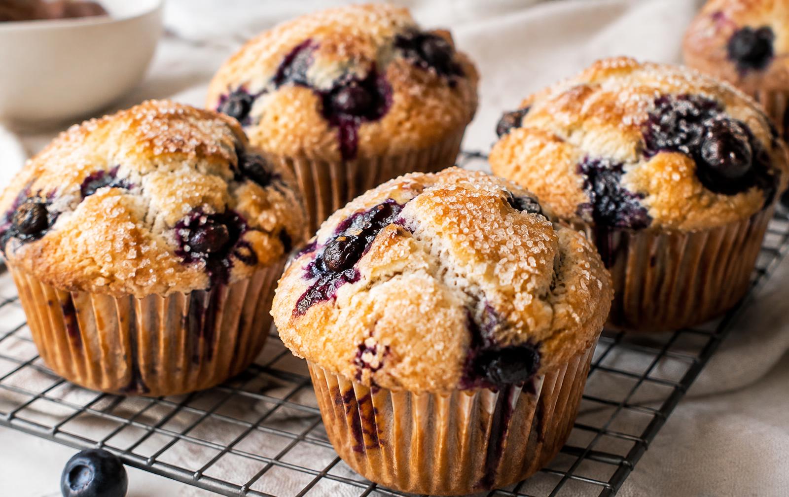 Tall bakery-style blueberry muffins with sparkling sugar tops and bursting blueberries, in paper liners on a wire rack