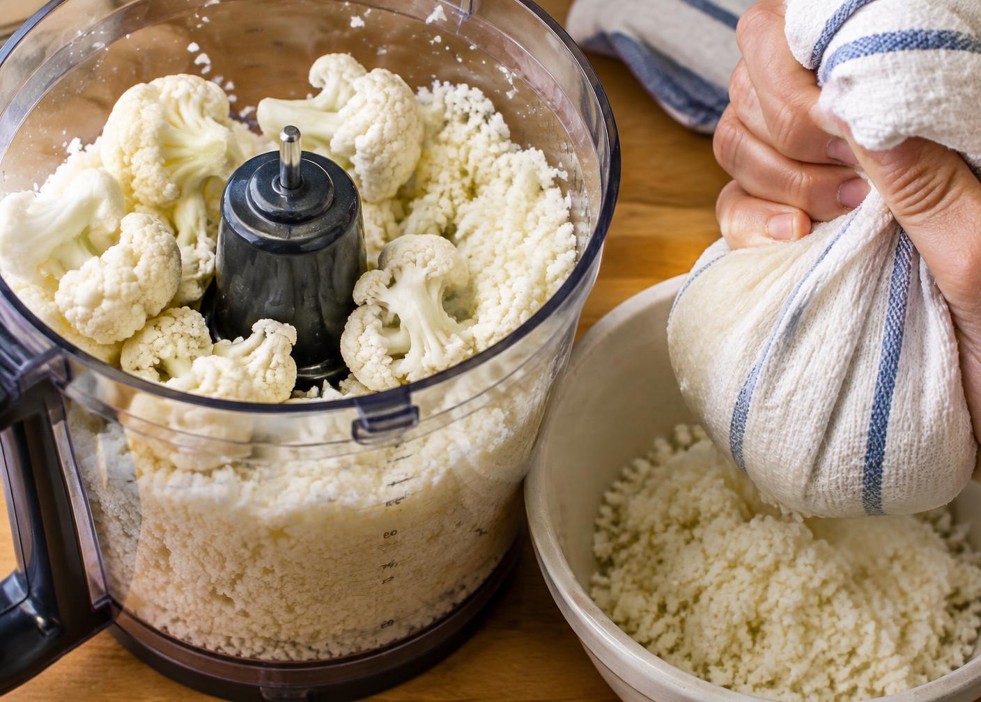 Cauliflower florets pulsed into rice-sized pieces in a food processor beside a towel being squeezed over a bowl of riced cauliflower