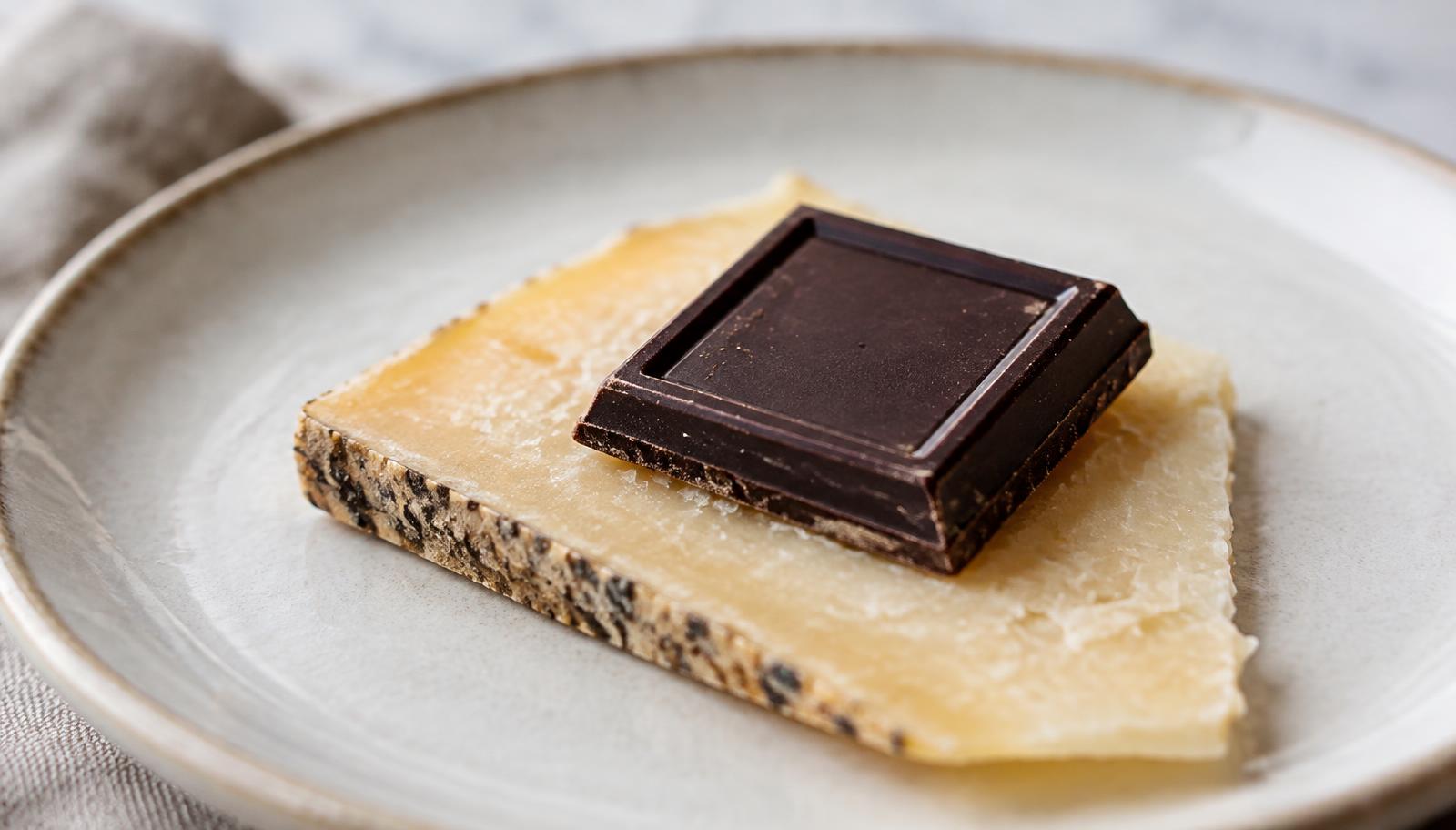 A square of dark chocolate resting on a thin slice of nutty aged alpine cheese on a pale ceramic plate