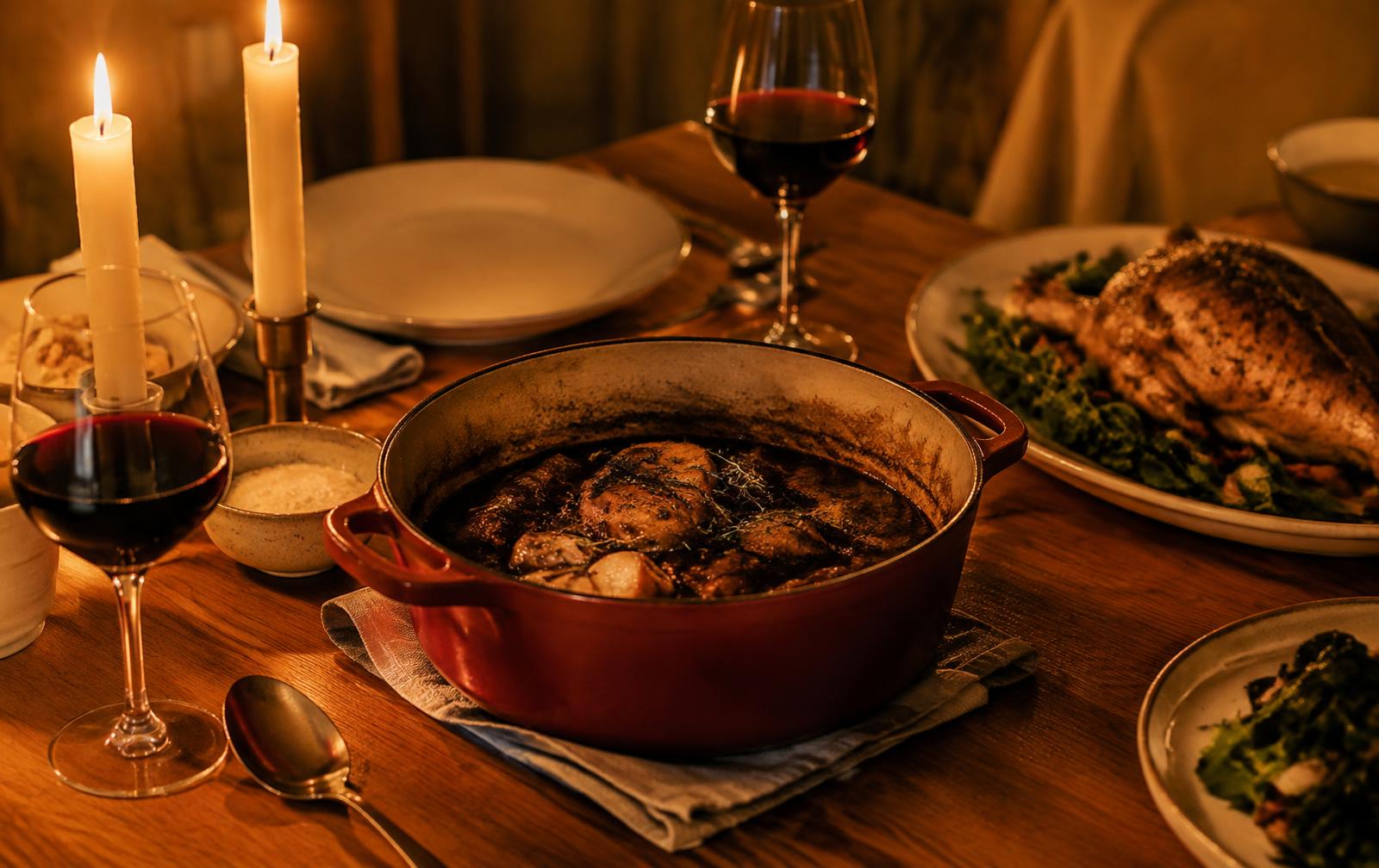 A candlelit dinner party table with a red Dutch oven of braised chicken, a roast platter and glasses of red wine