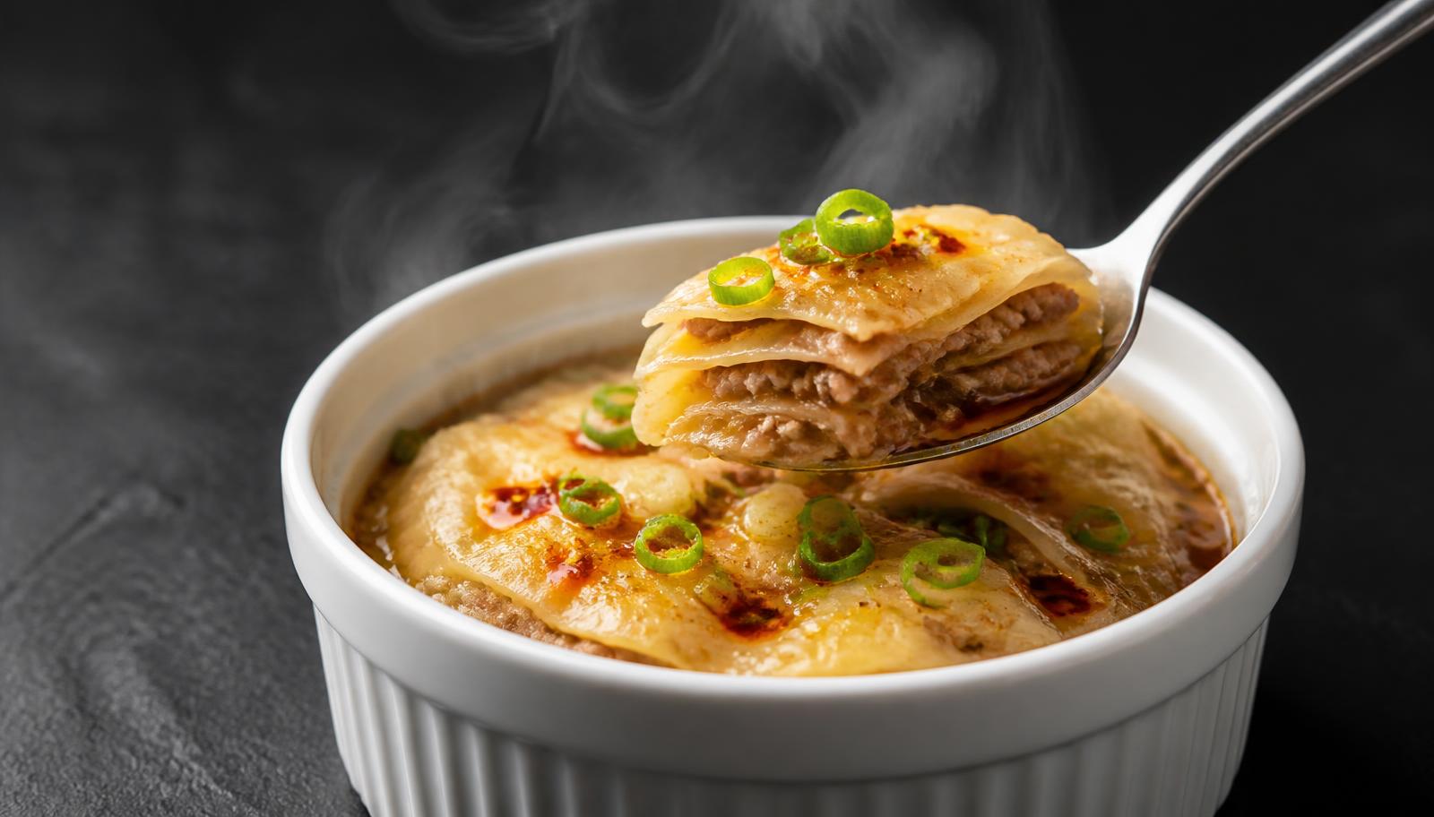A white ramekin of soup dumpling lasagna with layers of wonton wrapper and pork filling, scallions and chili oil, a spoon lifting one layered bite
