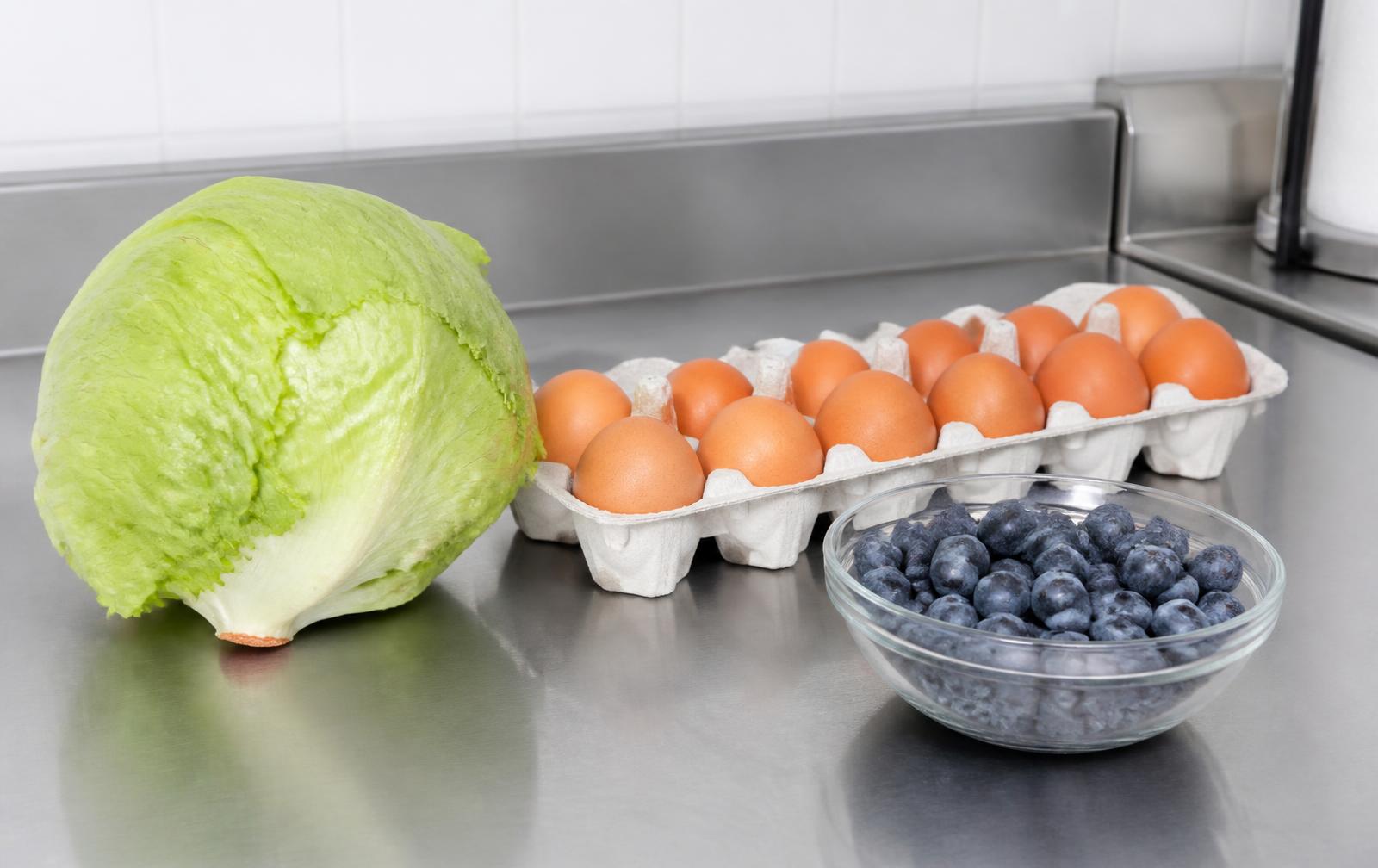 A head of iceberg lettuce, a carton of brown eggs, and a bowl of blueberries on a stainless steel counter