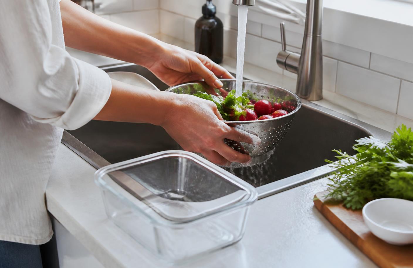 Hands rinsing fresh produce in a stainless steel colander under running water at a kitchen sink