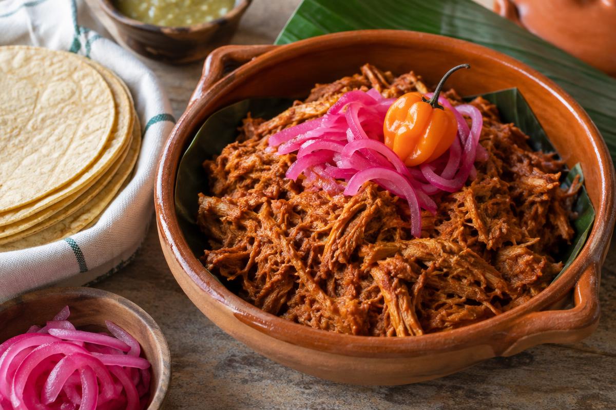 Shredded orange achiote-marinated cochinita pibil in a clay bowl with pickled red onion and a habanero