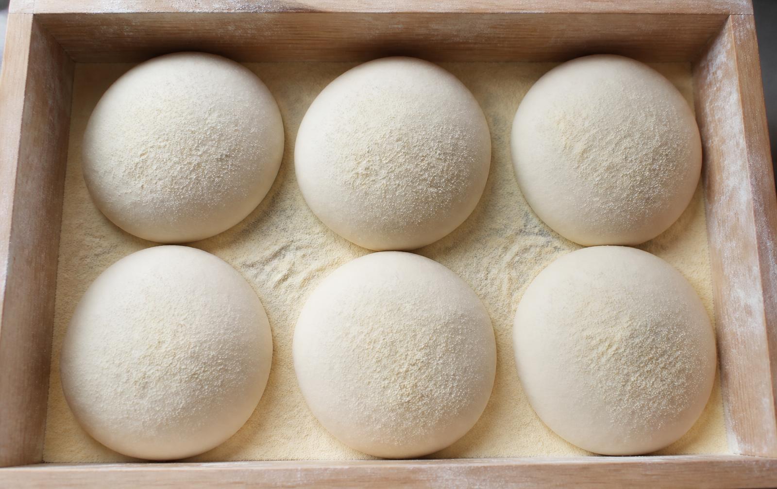 Six smooth round balls of pizza dough resting in a floured wooden proofing box dusted with semolina