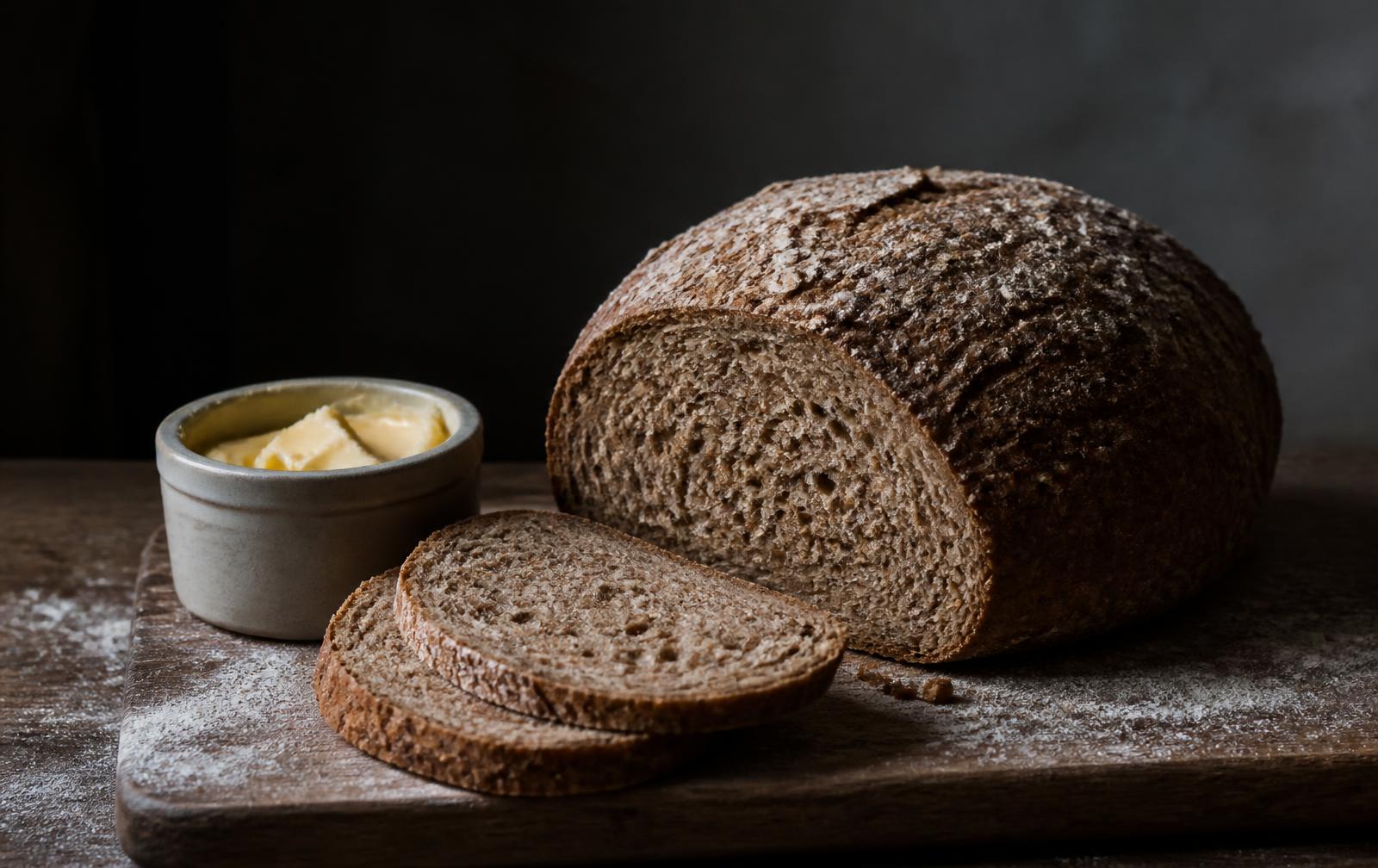 A dark coarse wholegrain loaf sliced open on a flour-dusted board beside a small crock of butter