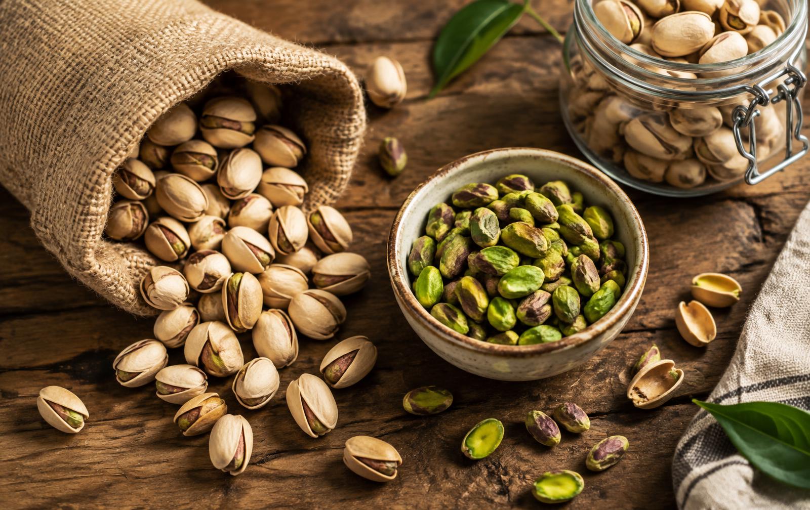 In-shell pistachios spilling from a burlap sack beside a bowl of green shelled kernels and a glass storage jar
