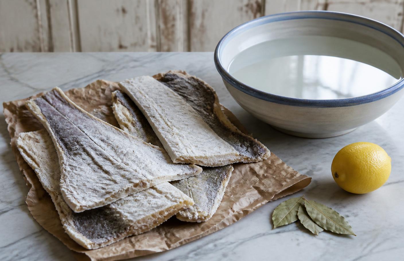 Dried salt cod fillets on butcher paper beside a bowl of soaking water, a lemon, and bay leaves