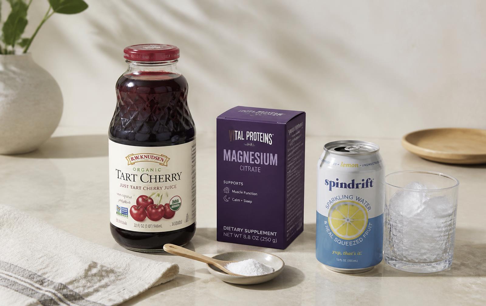 A bottle of tart cherry juice, a container of magnesium powder, a can of sparkling water and a glass of ice on a pale counter