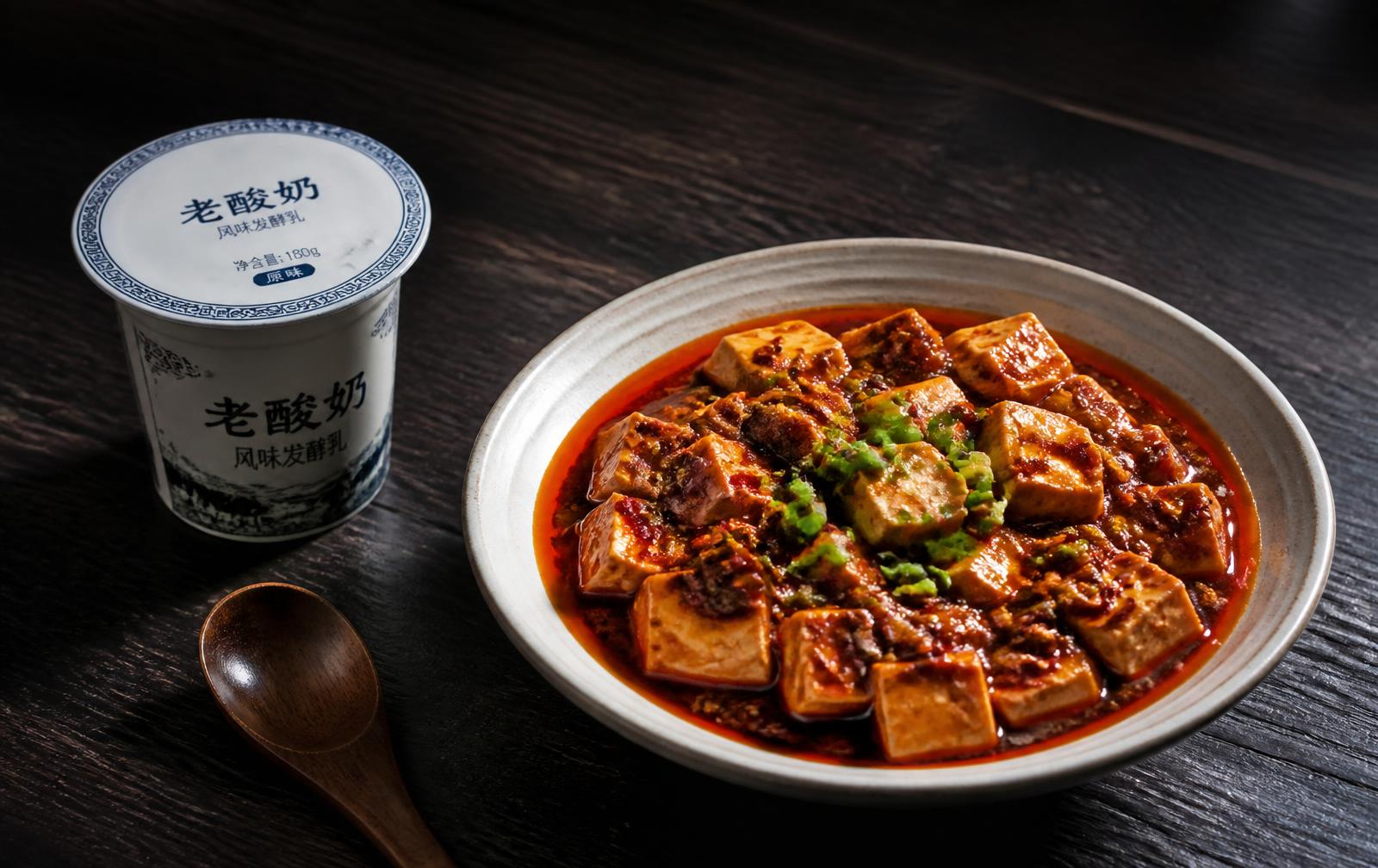 A cup of drinking yogurt beside a bowl of Sichuan mapo tofu in chili oil on a dark table