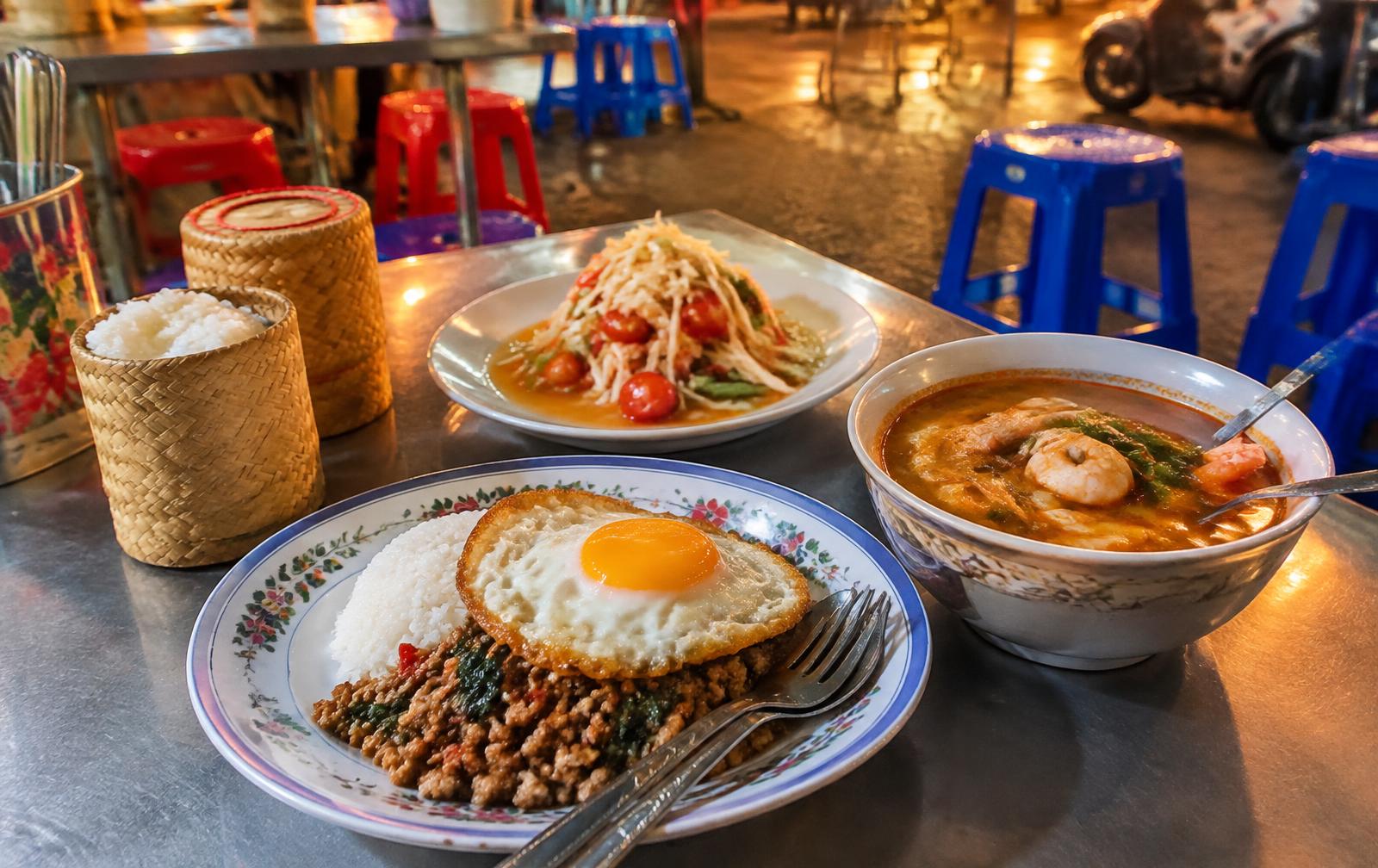 A Bangkok street food table with pad kra pao and a fried egg, som tam, tom yum soup and baskets of sticky rice beside plastic stools